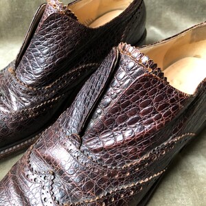 May include: A pair of brown leather shoes with a crocodile-embossed pattern. The shoes have a wingtip design and a lace-up closure.