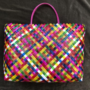 May include: A colorful woven tote bag with a pink handle. The bag is made of woven plastic strips in a variety of colors, including red, yellow, green, blue, and white. The bag is rectangular in shape.