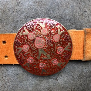 May include: A round, red and gold belt buckle with a floral design and stylised figures. The buckle is attached to a brown leather belt.