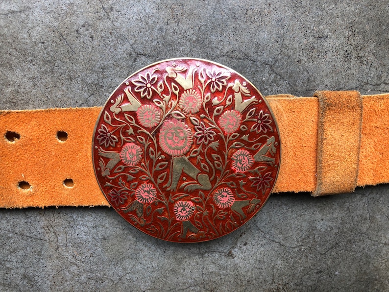 May include: A round, red and gold belt buckle with a floral design and stylised figures. The buckle is attached to a brown leather belt.