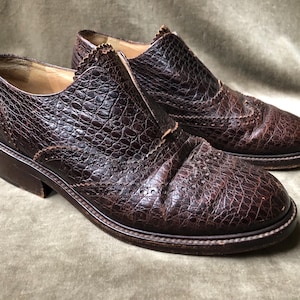 May include: A pair of brown leather shoes with a brogue design. The shoes have a textured, alligator-like pattern and a low heel.
