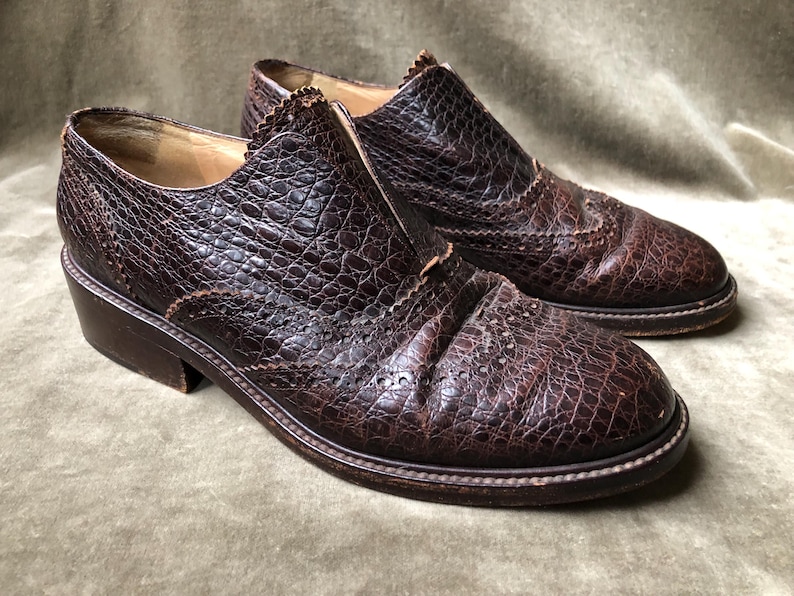May include: A pair of brown leather shoes with a brogue design. The shoes have a textured, alligator-like pattern and a low heel.
