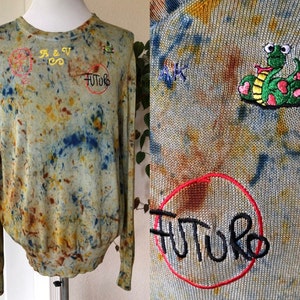 May include: A light green, blue, and brown tie-dye sweater with embroidered details. The sweater has a snake with hearts, the word "FUTURE" in a circle, and the letters "A & V" with a swirl.