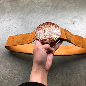 May include: A brown leather belt with a large, round, metal buckle. The buckle is decorated with a red and gold floral design.