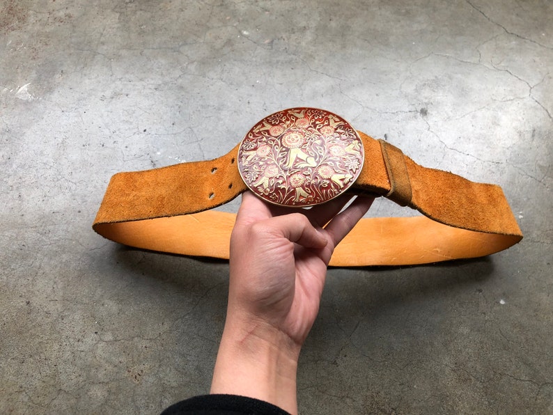 May include: A brown leather belt with a large, round, metal buckle. The buckle is decorated with a red and gold floral design.