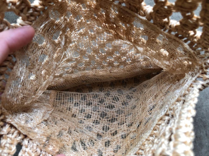 May include: Close-up of a beige crocheted bag with a pocket. The bag is made of a loosely woven, open-work material, creating a textured appearance. The pocket is visible inside the bag.