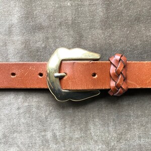 May include: A brown leather belt with a gold buckle and a braided leather strap detail.