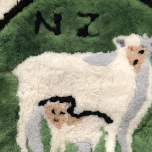 May include: A round, fluffy rug featuring a sheep design. The sheep is white with gray and tan accents, set against a green background with the letters "NZ" in black. The rug has a black border.