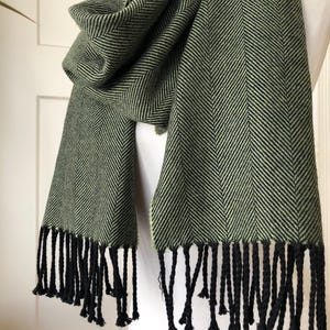 May include: A green and black herringbone pattern scarf with black fringe. The scarf is draped over a white form, showcasing the texture and design. The scarf's color palette is a mix of olive green and black.