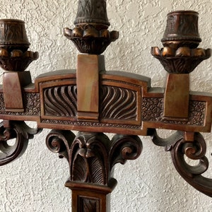Puede incluir: Un candelabro de madera con cinco portavelas, con intrincados tallados y un acabado marrón oscuro. El candelabro tiene una parte superior decorativa con un diseño curvo y un pilar central.