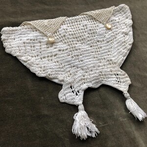 Può includere: Una borsetta bianca all'uncinetto dalla forma unica. La borsetta presenta due piccoli lembi triangolari con bottoni perlati e due nappe bianche. Il design traforato e i dettagli delicati suggeriscono un oggetto vintage o fatto a mano.