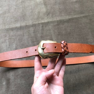 May include: A brown leather belt with a gold buckle and a braided leather detail. The belt is shown on a brown fabric background.