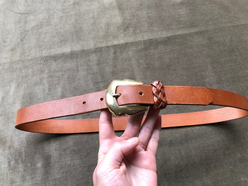 May include: A brown leather belt with a gold buckle and a braided leather detail. The belt is shown on a brown fabric background.