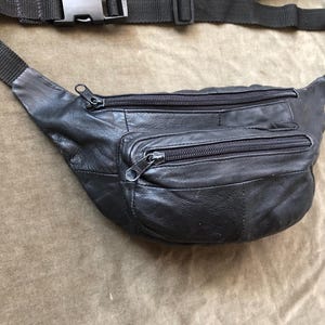 May include: Black leather fanny pack with two zippered compartments and a smaller front pocket. The adjustable black strap has a plastic buckle. The fanny pack is designed to be worn around the waist or across the body.