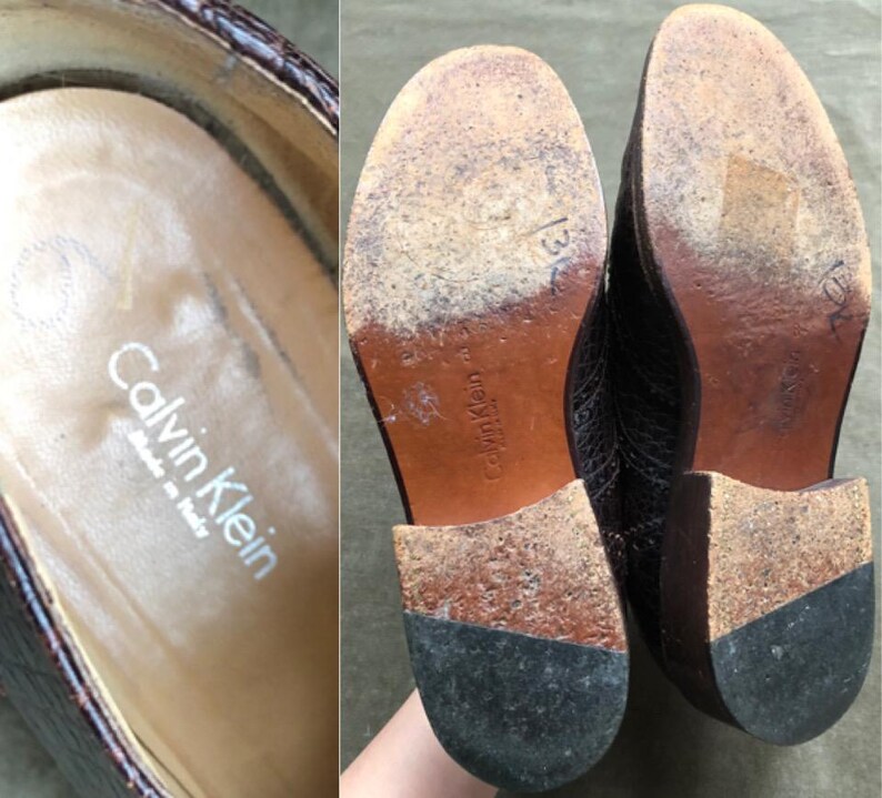 May include: A pair of brown leather shoes with a crocodile pattern. The shoes have a black rubber sole and the text "Calvin Klein" is printed on the inside of the shoe.