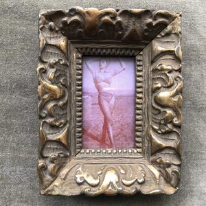 May include: A rectangular picture frame with a heavy, ornate, metallic gold finish featuring deep relief carvings. The frame holds a small, vertical photograph of a person in a two-piece swimsuit standing on a beach with a reddish tint.