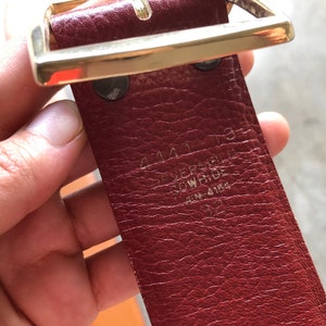 May include: A close-up of a red leather belt with a gold buckle. The belt has the text "44413 VERSACE" and "ITEM 4184" embossed on the back.