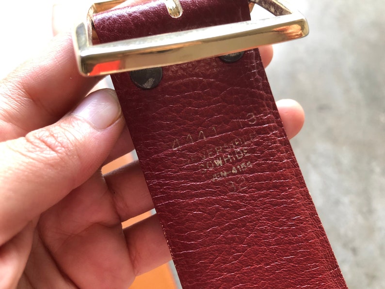 May include: A close-up of a red leather belt with a gold buckle. The belt has the text "44413 VERSACE" and "ITEM 4184" embossed on the back.