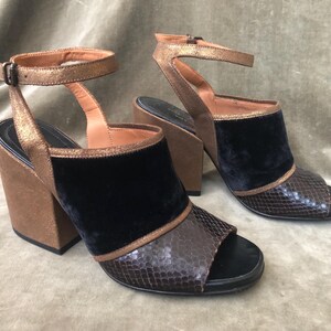 May include: A pair of open-toe heeled sandals. The shoes feature a black velvet vamp, a snakeskin-textured toe, and a metallic bronze ankle strap. The chunky heel and platform are brown suede. The sandals have a buckle closure.