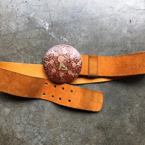 May include: A brown leather belt with a round, ornate metal buckle. The buckle features a red and gold design with floral and geometric patterns.