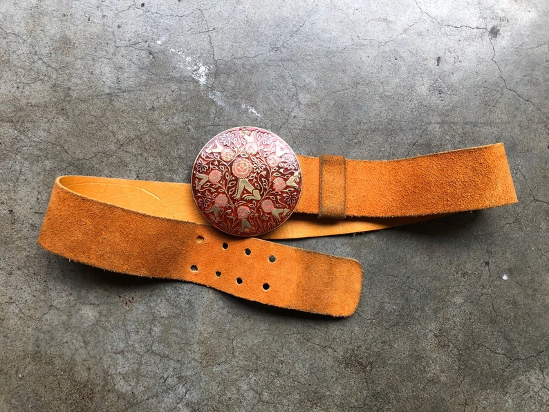 May include: A brown leather belt with a round, ornate metal buckle. The buckle features a red and gold design with floral and geometric patterns.