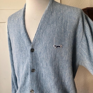 May include: A light blue V-neck cardigan sweater with a textured knit finish. The garment features three dark buttons down the front placket and a small embroidered patch on the left chest depicting a dark blue fox in a running pose.