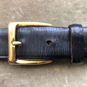 May include: Close-up of a black leather belt with a gold-colored rectangular buckle. The belt shows signs of wear, with visible creases and a worn appearance. The buckle is shiny and contrasts with the matte finish of the belt.