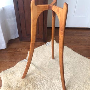 May include: A tall, three-legged wooden plant stand crafted from light brown wood with a smooth, polished finish. The stand features curved, slender legs that extend upward to form a central support structure with three distinct, upward-facing prongs.