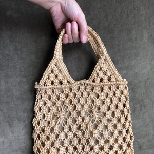 May include: A beige macrame tote bag with a triangular handle and open weave design. The bag is held by a hand against a dark green background. The bag's open weave pattern features floral-like details.