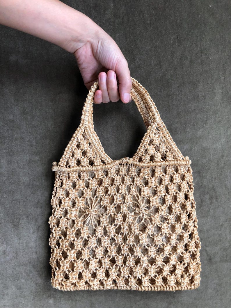 May include: A beige macrame tote bag with a triangular handle and open weave design. The bag is held by a hand against a dark green background. The bag's open weave pattern features floral-like details.