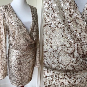 May include: A long-sleeved, sequined dress with a V-neckline and a wrap-style bodice. The dress is a light gold color with brown sequins.