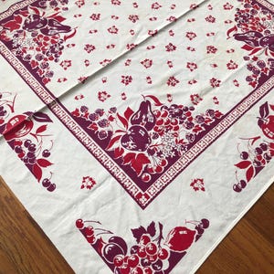 May include: A white tablecloth with a repeating pattern of red and purple fruit and floral designs. The border features a geometric pattern. The tablecloth is folded on a wooden surface.