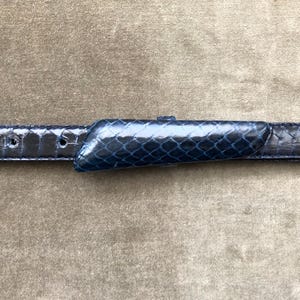 May include: A navy blue belt with a textured, snakeskin-like pattern. The belt features a matching buckle with a similar design. The belt has several holes for adjustment.