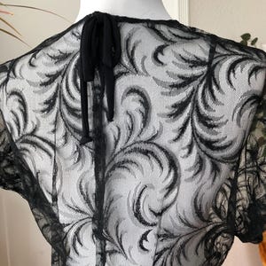 May include: Black lace top with a delicate floral pattern. The short-sleeved top features a tie closure at the back of the neck. The intricate design is visible against a white background, showcasing the detailed craftsmanship.