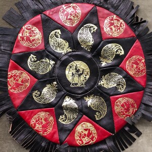 Può includere: Elemento decorativo rotondo in pelle, con un motivo centrale a stella. Segmenti rossi e neri alternati con motivi dorati a paisley e animali. Il cerchio centrale raffigura un cammello e un cavaliere. Frange nere sul bordo.