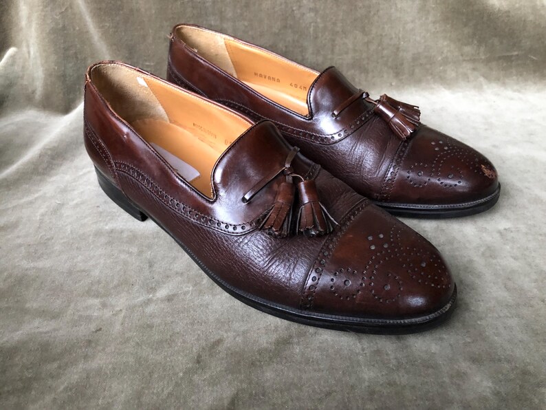 May include: A pair of brown leather tassel loafers with a wingtip design and a perforated pattern. The loafers have a brown leather tassel on each shoe.