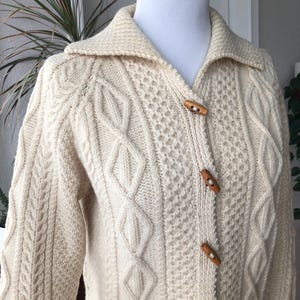 May include: Cream-colored, cable-knit cardigan sweater with a collared neckline and wooden toggle closures. The sweater features intricate knit patterns and long sleeves, displayed on a mannequin.