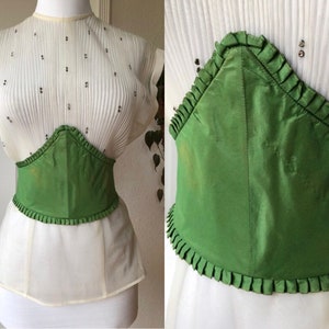 May include: A green satin corset with a ruffled trim. The corset is cinched at the waist and has a pointed shape in the front.