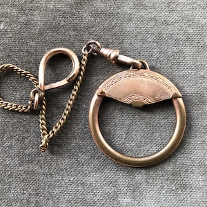 May include: A rose gold-toned metal watch fob featuring a circular ring attached to a decorative fan-shaped top with engraved line patterns. The piece includes a short, delicate curb chain connected to a swivel clasp and a small, looped metal hook.