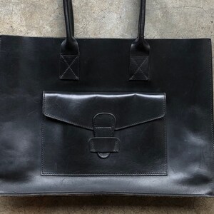 Puede incluir: Un bolso tote de cuero negro con un bolsillo frontal grande y un asa superior.