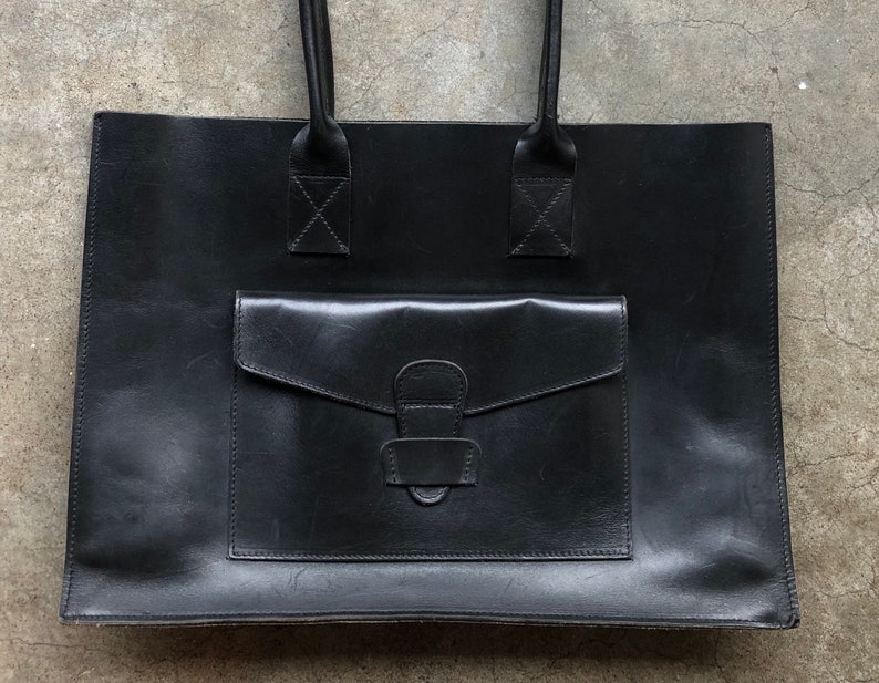 Puede incluir: Un bolso tote de cuero negro con un bolsillo frontal grande y un asa superior.