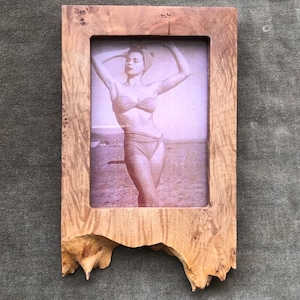 May include: A rectangular wooden picture frame with a natural burl wood grain pattern and an irregular, raw-edged bottom. The frame contains a sepia-toned photograph of a person in a two-piece swimsuit posing with arms raised above their head.