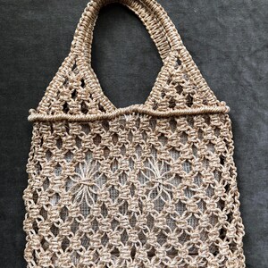 May include: A beige macrame tote bag with two handles. The bag features a diamond-patterned weave with a floral design in the center. The bag is made of a natural fiber material and is suitable for carrying items.