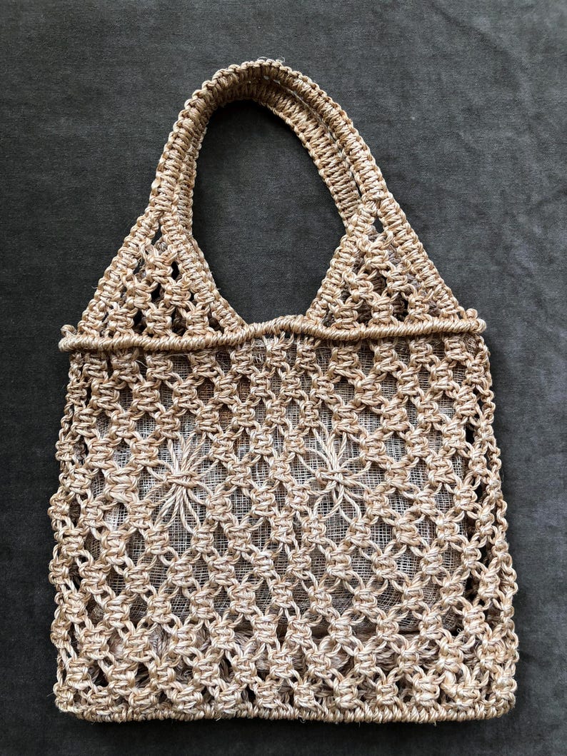 May include: A beige macrame tote bag with two handles. The bag features a diamond-patterned weave with a floral design in the center. The bag is made of a natural fiber material and is suitable for carrying items.