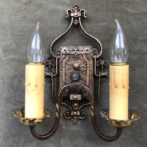 May include: Ornate, antique wall sconce with two faux candle lights. The sconce features a decorative metal frame in bronze and gold tones, with a central panel and curved arms holding the candle-shaped lights. The bulbs are clear, and the sconce is mounted on a gray wall.