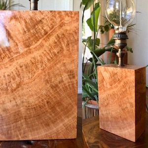 Puede incluir: Una base de lámpara de madera con un patrón de veta de madera natural. La base es rectangular y tiene un color marrón claro. La lámpara está coronada con un globo de vidrio transparente y una lámpara de latón.