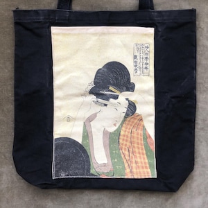 May include: Black tote bag featuring a Japanese woodblock print of a woman applying makeup. The print is in shades of green, orange, and beige, with Japanese characters. The bag has black handles and is made of a durable fabric.