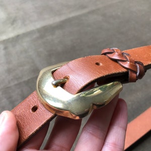 May include: A brown leather belt with a braided leather detail and a gold metal buckle.