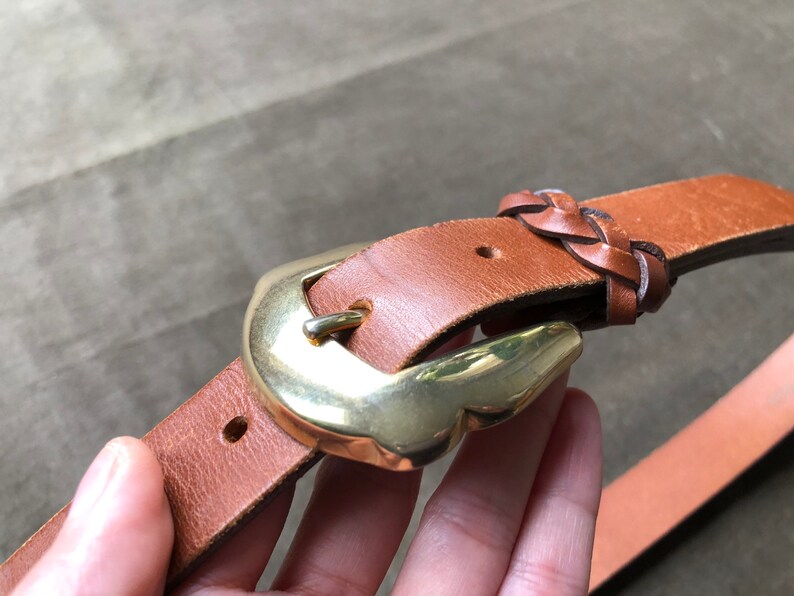 May include: A brown leather belt with a braided leather detail and a gold metal buckle.