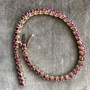 Puede incluir: Una pulsera de tenis en tono dorado con una hilera continua de piedras preciosas redondas de color rosa intenso engastadas en garras metálicas individuales. La pulsera incluye un cierre de caja metálico seguro con un pestillo de seguridad.